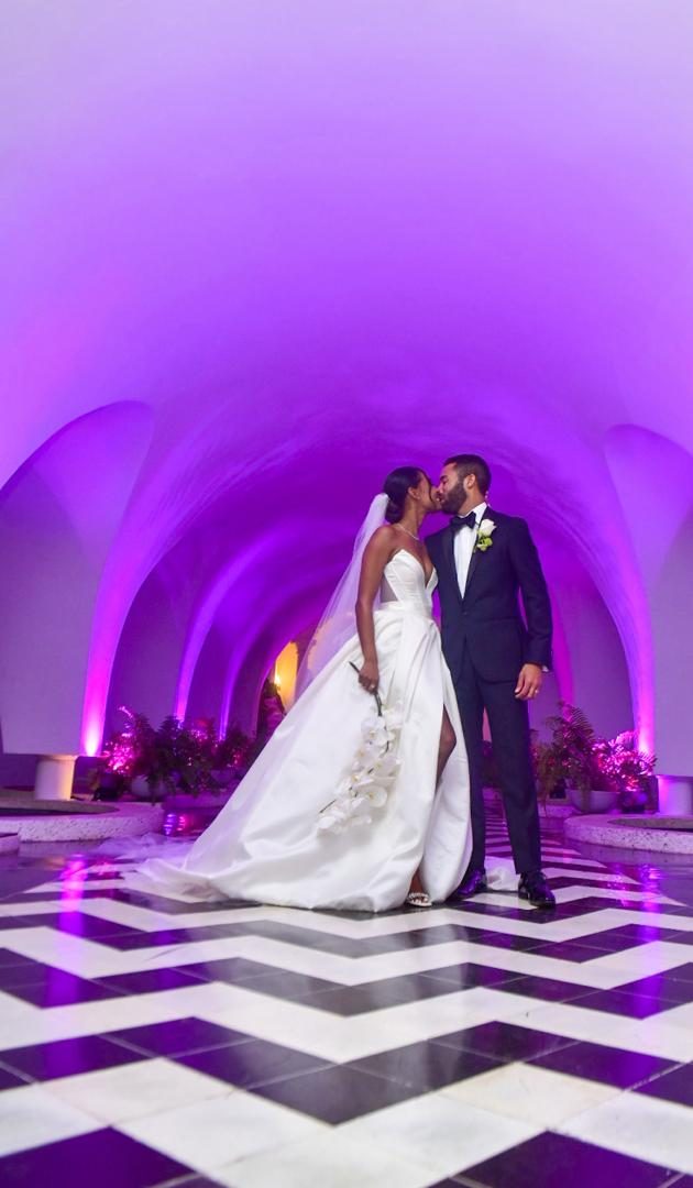 Bride and groom kissing beneath a violet-lit vaulted arcade